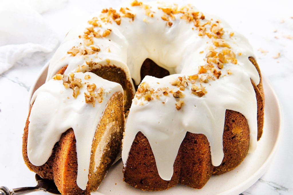 A round carrot bundt cake with cream cheese filling and white icing drizzled over the top, garnished with chopped nuts, sits on a white plate against a light background.