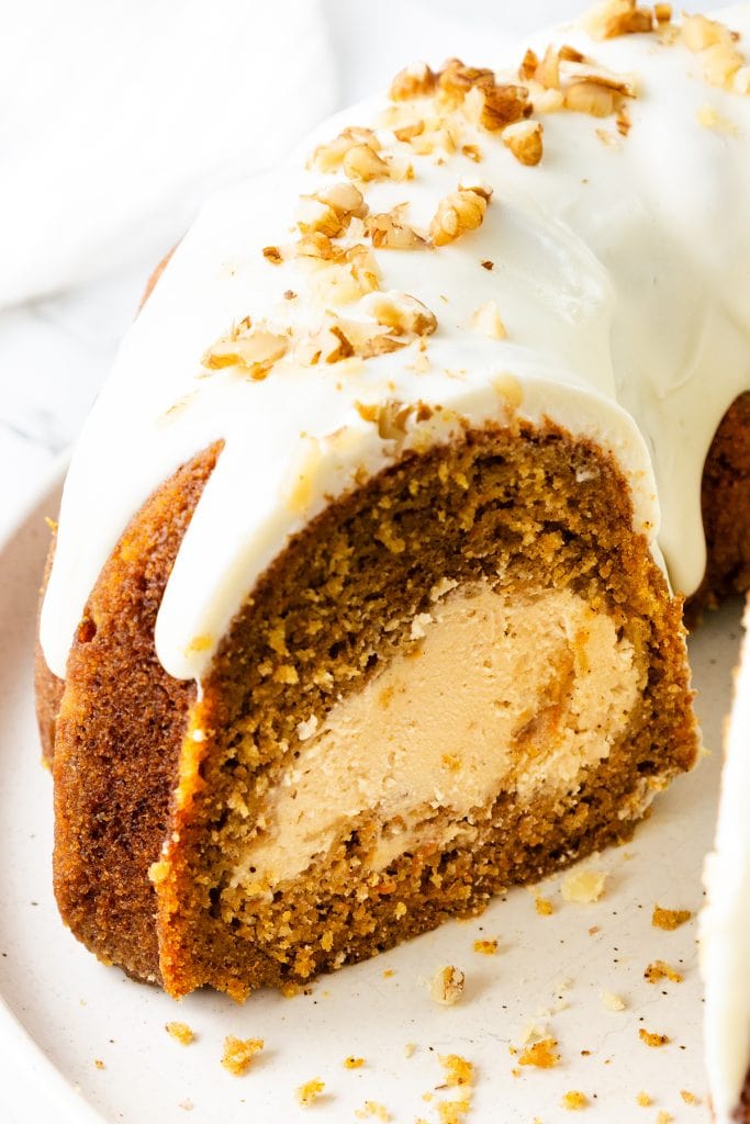 A close-up of a sliced Bundt Cake with a luscious cream cheese filling, topped with white icing and chopped nuts, displayed on a plate.