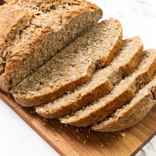A loaf of Irish Soda Bread, partially sliced, sits on a wooden cutting board on a white marble surface.