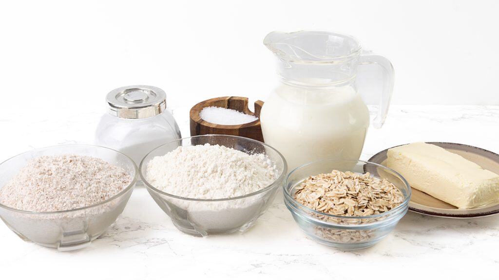 A selection of baking ingredients for Irish Soda Bread on a white surface, including a glass pitcher of milk, bowls of sugar, flour, oats, whole wheat flour, a plate of butter, and a small container of salt.