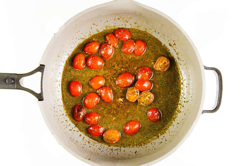 A white pan with a black handle contains red cherry tomatoes cooking in a greenish-brown sauce, perfect for Italian Smothered Chicken, viewed from above.