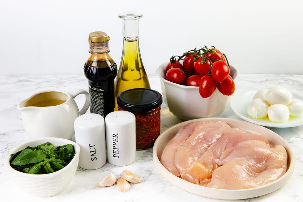  raw chicken breasts, grape tomatoes, mozzarella balls, fresh basil, garlic cloves, seasonings, a jar of pesto, oil, vinegar, and a small pitcher of broth.