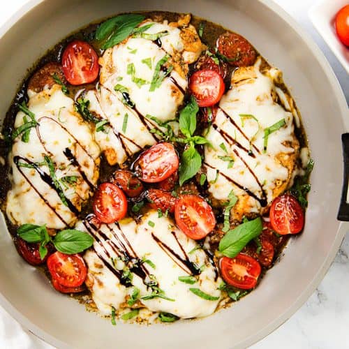 A skillet with Italian Smothered Chicken breasts topped with melted mozzarella, cherry tomatoes, fresh basil, and a drizzle of balsamic glaze. A small bowl of cherry tomatoes sits nearby on a white surface.
