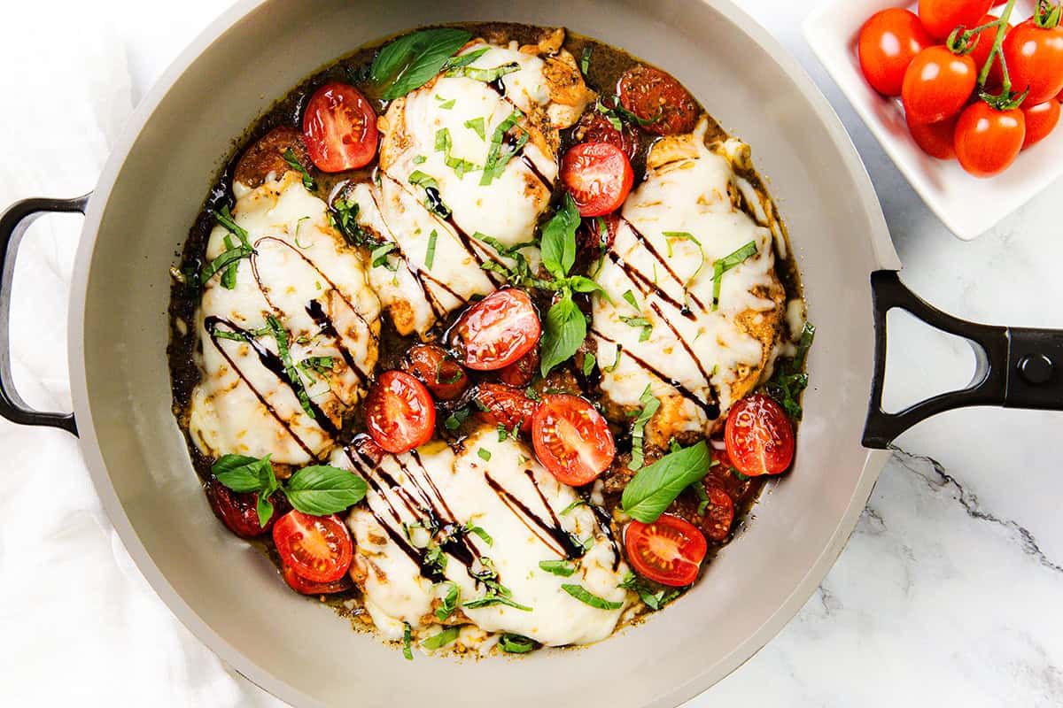 A skillet with Italian Smothered Chicken breasts topped with melted mozzarella, cherry tomatoes, fresh basil, and a drizzle of balsamic glaze. A small bowl of cherry tomatoes sits nearby on a white surface.