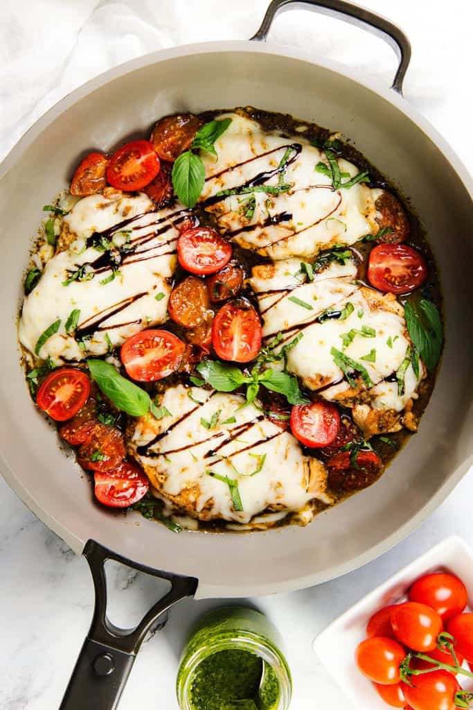 A skillet filled with Italian Smothered Chicken—cooked chicken breasts topped with melted cheese, cherry tomatoes, fresh basil, and balsamic glaze—served alongside a jar of pesto and a cluster of tomatoes.