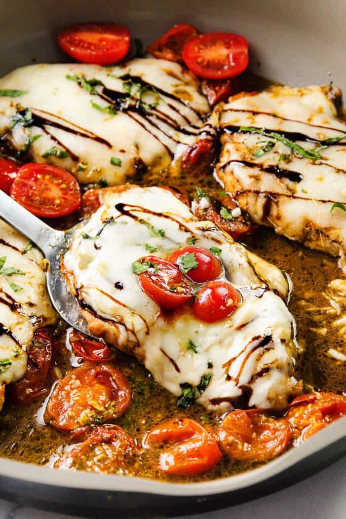 Italian Smothered Chicken breasts topped with melted cheese, cherry tomatoes, chopped herbs, and a drizzle of balsamic glaze are cooked in a skillet with a savory sauce. A spatula lifts one piece, showcasing this juicy, flavorful dish.