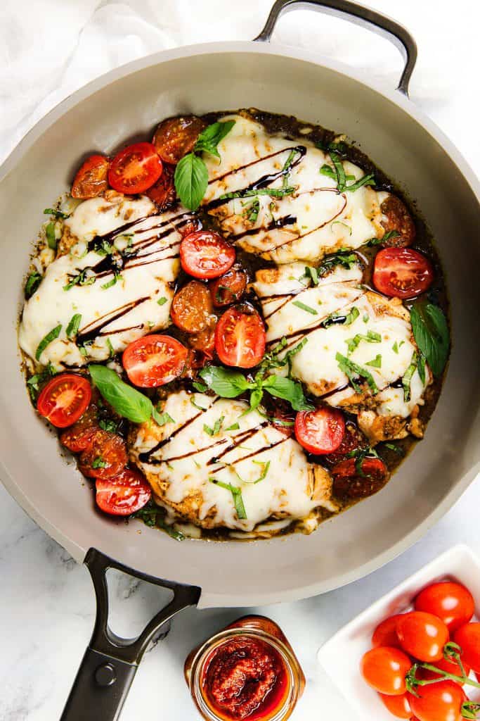 A skillet filled with Italian Smothered Chicken—tender chicken breasts topped with melted cheese, fresh basil, and balsamic glaze, surrounded by cooked cherry tomatoes and garnished with basil. Fresh and sun-dried tomatoes sit nearby.