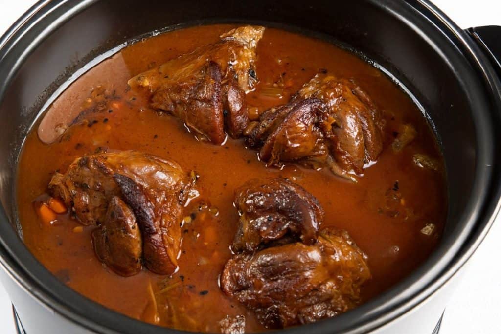 Lamb shanks in a rich, reddish-brown sauce inside a black slow cooker or pot. 