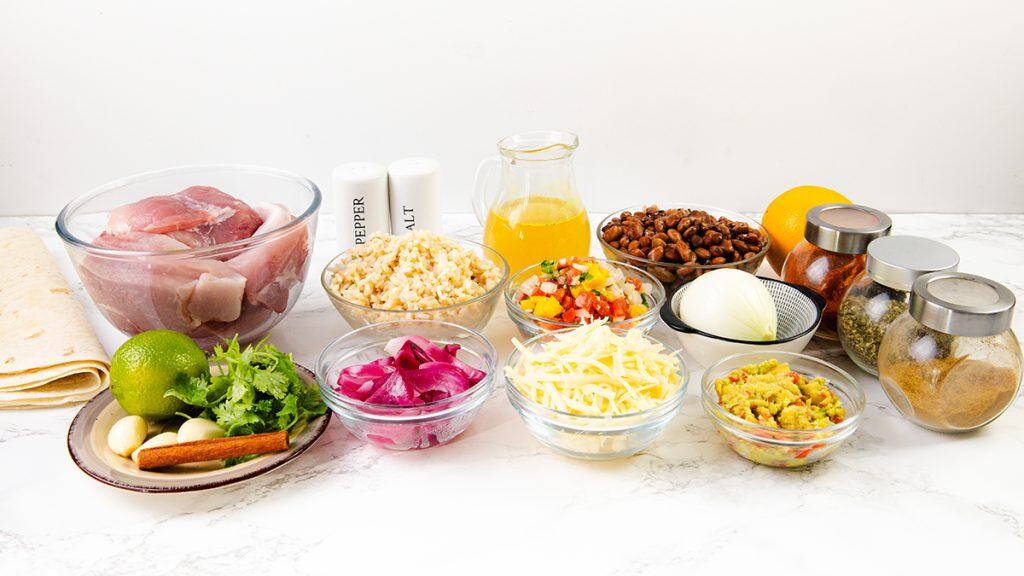 An assortment of ingredients for a Mexican-inspired dish, perfect for burritos, including raw chicken, tortillas, cheese, rice, beans, onion, lime, cilantro, spices, garlic, salsa, guacamole, broth, and pickled onions arranged on a white surface.