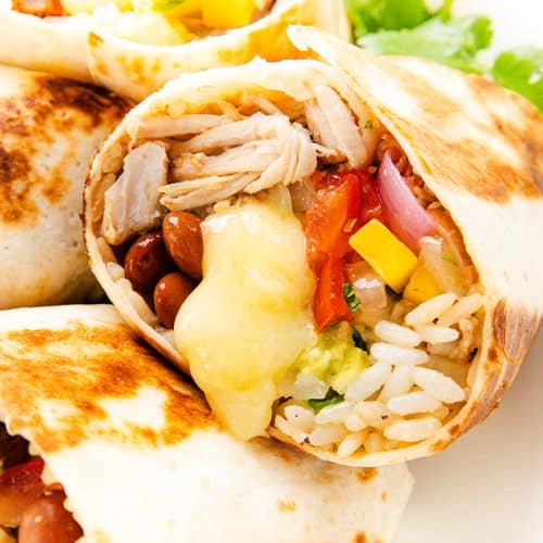 A close-up of a burrito cut in half, showing fillings of slow cooker carnitas, melted cheese, beans, rice, guacamole, diced tomatoes, and onions all wrapped in a grilled tortilla&mdash;perfect for burritos lovers.