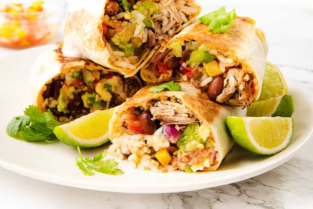 Four burrito halves filled with slow cooker carnitas, rice, beans, guacamole, and vegetables are arranged on a white plate, garnished with cilantro and lime wedges.