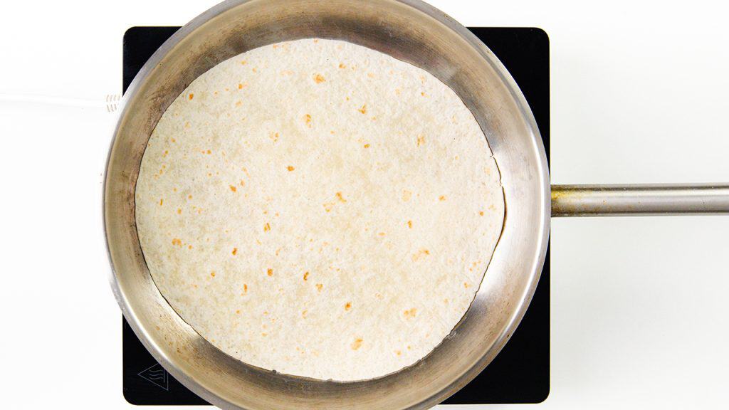 A flour tortilla, perfect for burritos, is heating in a stainless steel pan on a stovetop, viewed from above. The tortilla has light brown spots and the pan handle extends to the right.