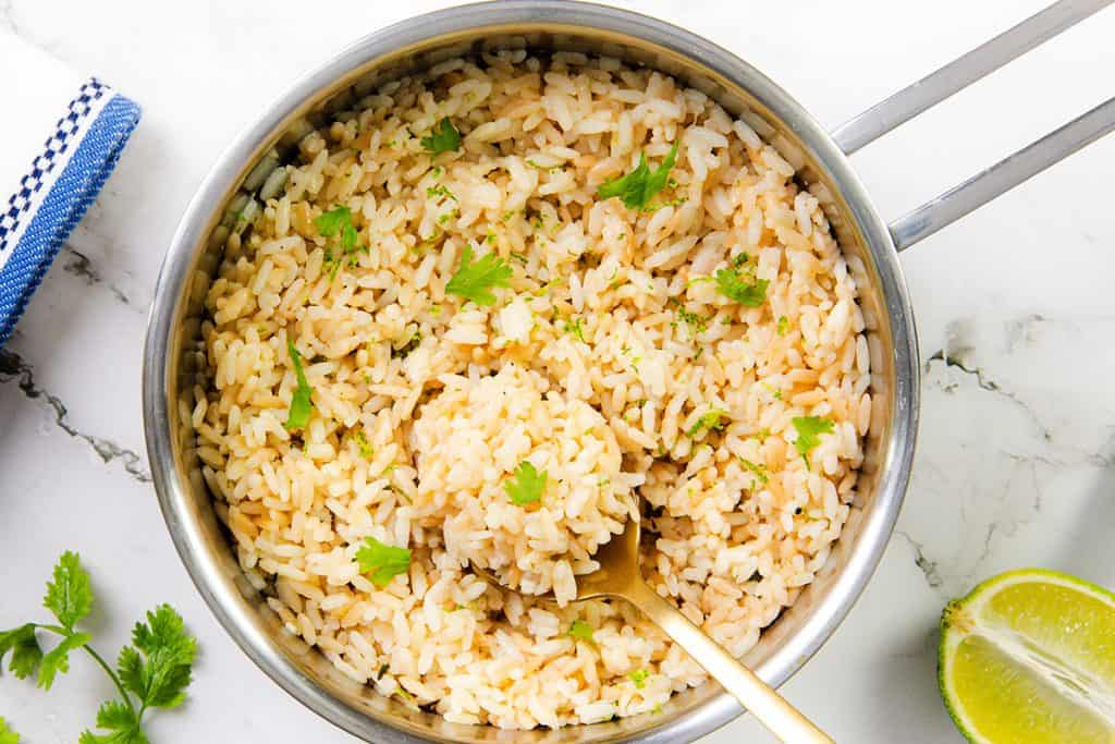 A pot of cilantro rice garnished with chopped cilantro, with a gold spoon resting inside. A lime wedge and a sprig of cilantro are nearby on a marble surface.