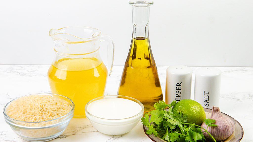 A pitcher of broth, a bottle of oil, dry rice for Cilantro Lime Rice, a bowl of cream for extra Creamy Rice, salt and pepper shakers, a lime, cilantro, and garlic cloves arranged on a white surface with a white background.