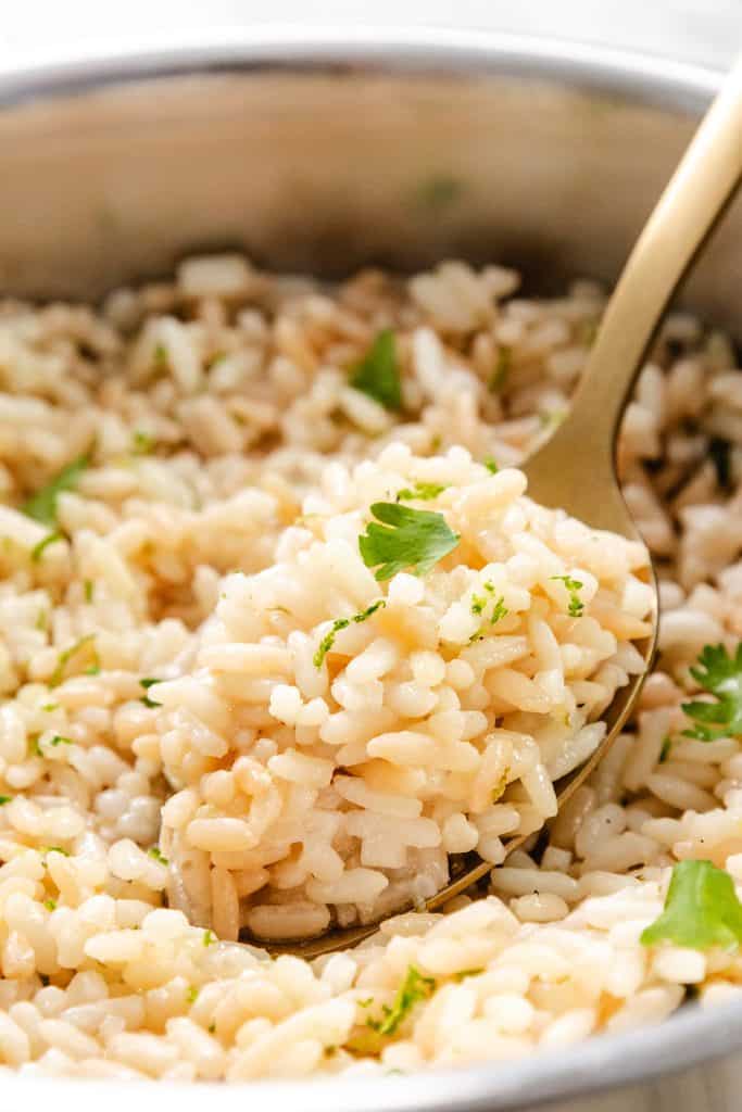 A close-up of a gold-colored spoon scooping fluffy, cooked cilantro lime rice garnished with fresh parsley from a pot. The rice appears tender and fragrant.