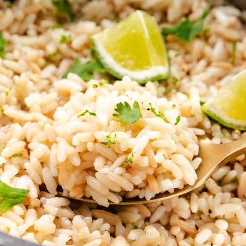 A close-up of cilantro lime rice garnished with fresh cilantro and lime wedges. A gold spoon holds a portion, showcasing visible zest and herbs throughout.