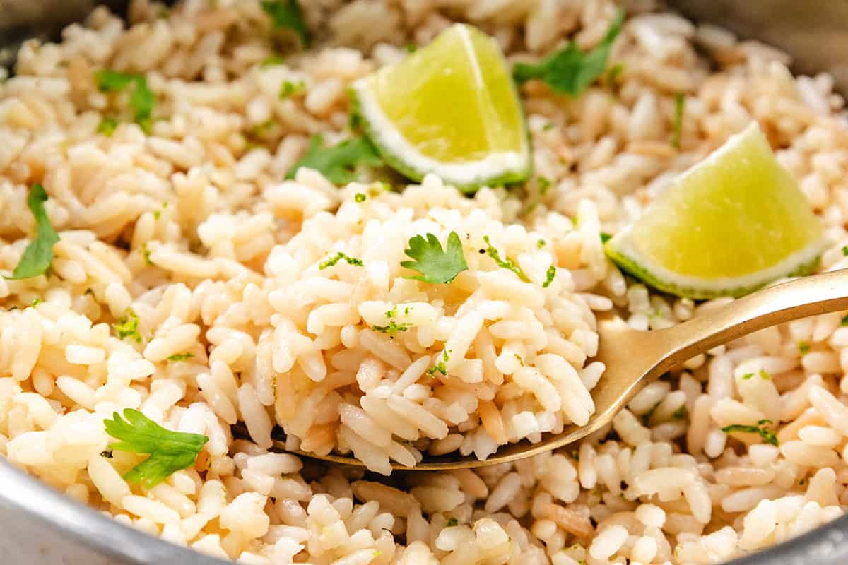 A close-up of cilantro lime rice garnished with fresh cilantro and lime wedges. A gold spoon holds a portion, showcasing visible zest and herbs throughout.