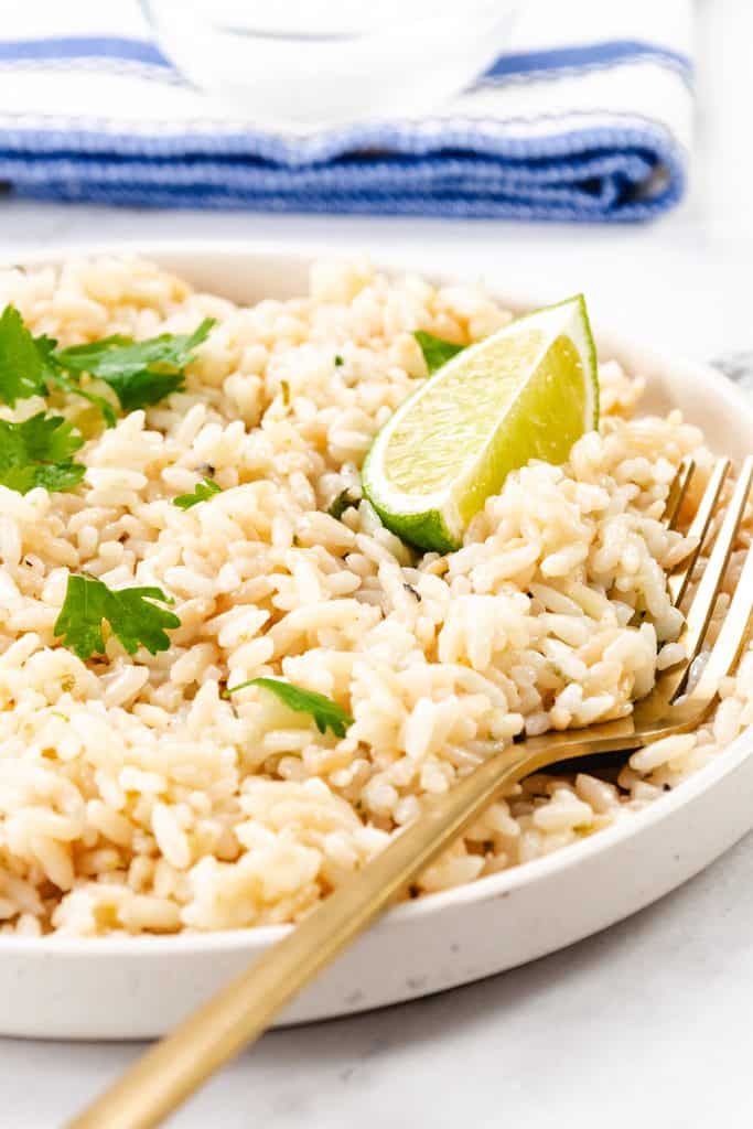 A plate of cilantro lime rice garnished with fresh cilantro and a lime wedge, with a gold fork resting on the side. A blue and white striped napkin is in the background.