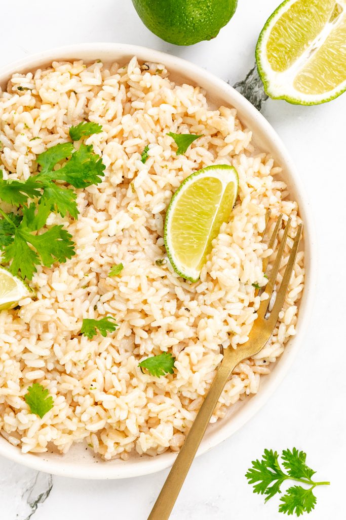 A bowl of creamy cilantro lime rice garnished with fresh cilantro and lime wedges, with a gold fork resting on the side. A halved lime and cilantro leaves are placed nearby on a white surface.