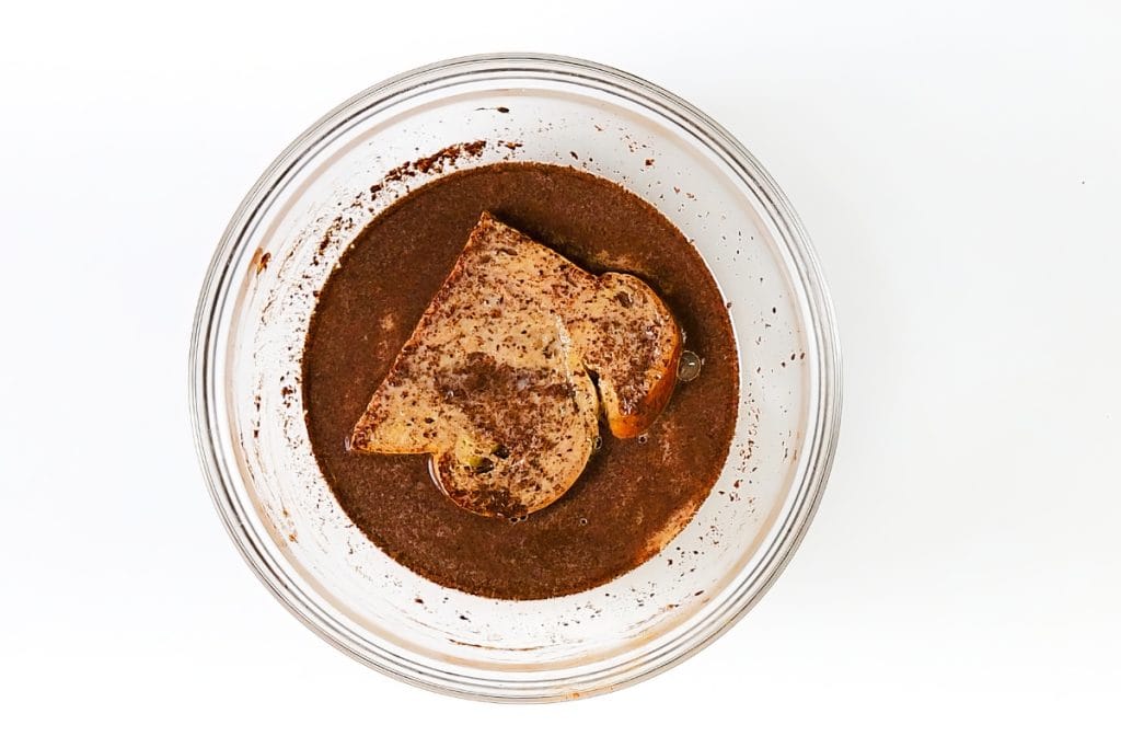 A glass bowl containing a brown, liquid mixture with two slices of bread soaking in it, viewed from above on a white background—perfect for making classic French Toast.