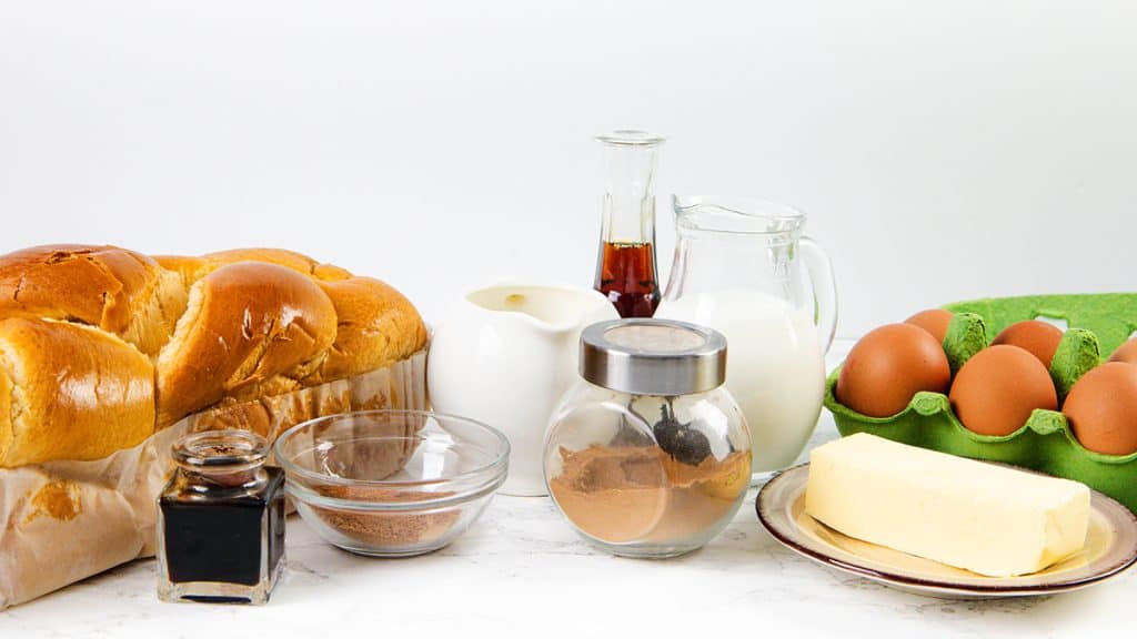A loaf of brioche bread, a jar of vanilla, a small pitcher of cream, eggs, butter, cinnamon, and brown sugar—all the classic ingredients you need to make delicious French Toast—are beautifully arranged on a white background.