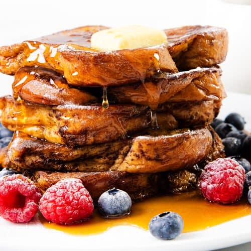 A delicious stack of French toast topped with butter and syrup, surrounded by fresh raspberries and blueberries on a white plate.