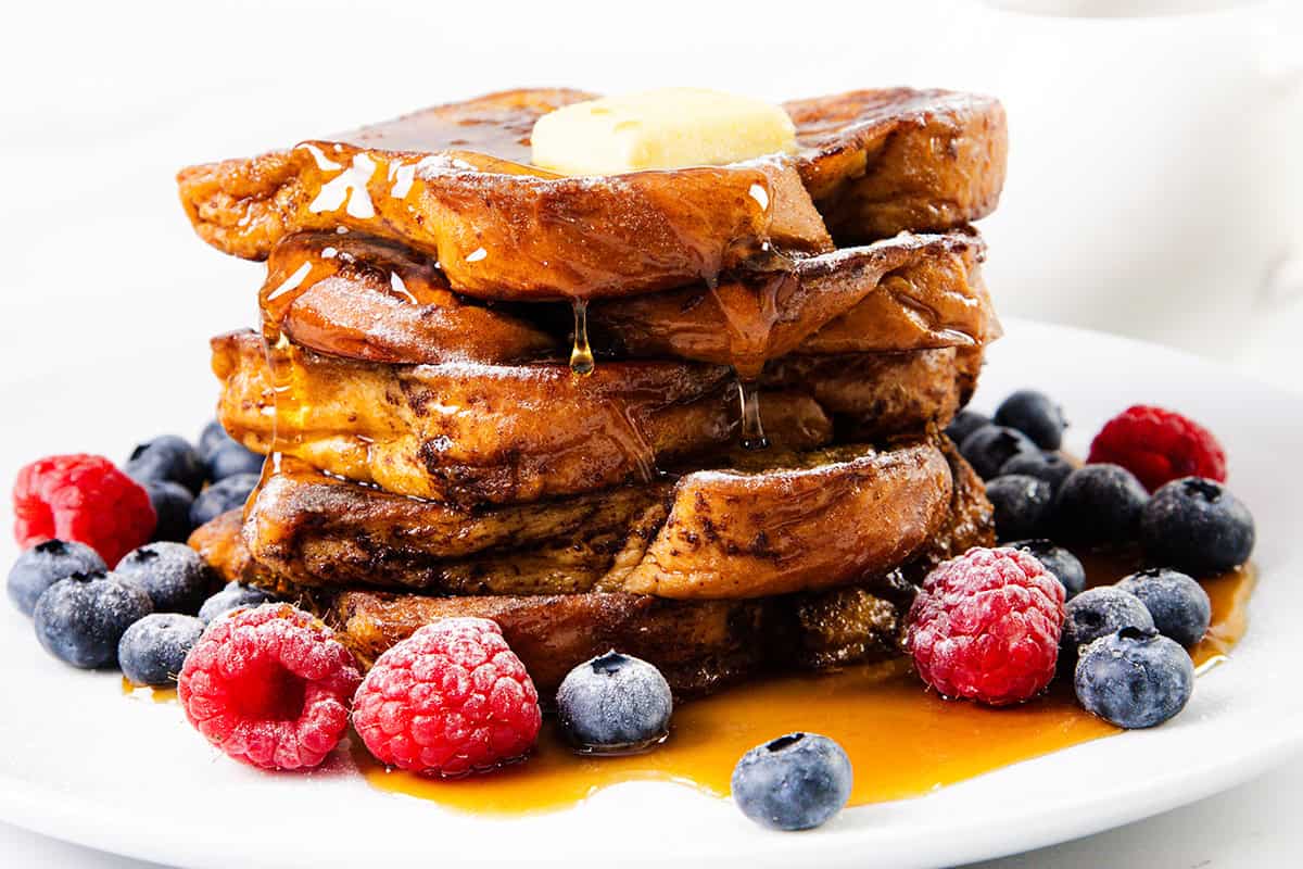 A delicious stack of French toast topped with butter and syrup, surrounded by fresh raspberries and blueberries on a white plate.