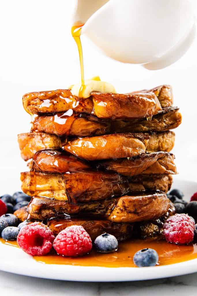 A stack of delicious French Toast topped with butter and syrup, surrounded by fresh blueberries and raspberries, with golden syrup being poured from a white pitcher above.