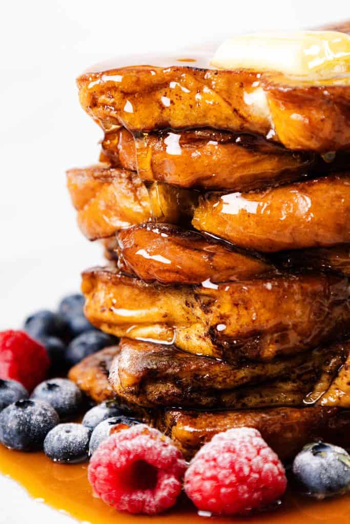 A stack of golden pancakes topped with a pat of butter and drizzled with syrup, surrounded by fresh blueberries and raspberries on a white plate—perfect for any breakfast spread alongside classic French Toast.