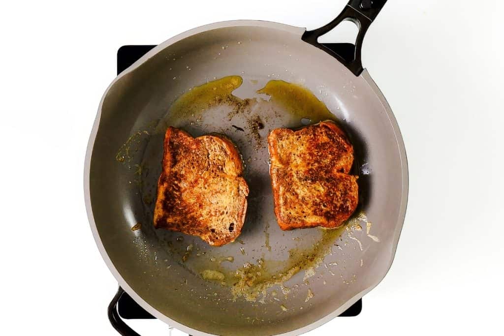 A gray frying pan with two slices of French toast sizzling in oil, shown from above against a white background.
