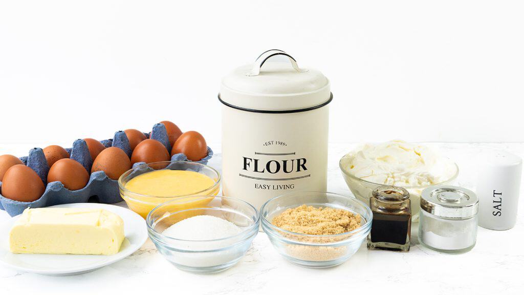 A variety of baking ingredients on a white surface, including eggs, flour, butter, sugar, brown sugar, cream cheese for Lemon Cheesecake Bars, salt, vanilla extract, and a bowl of yellow mixture&mdash;all arranged neatly.