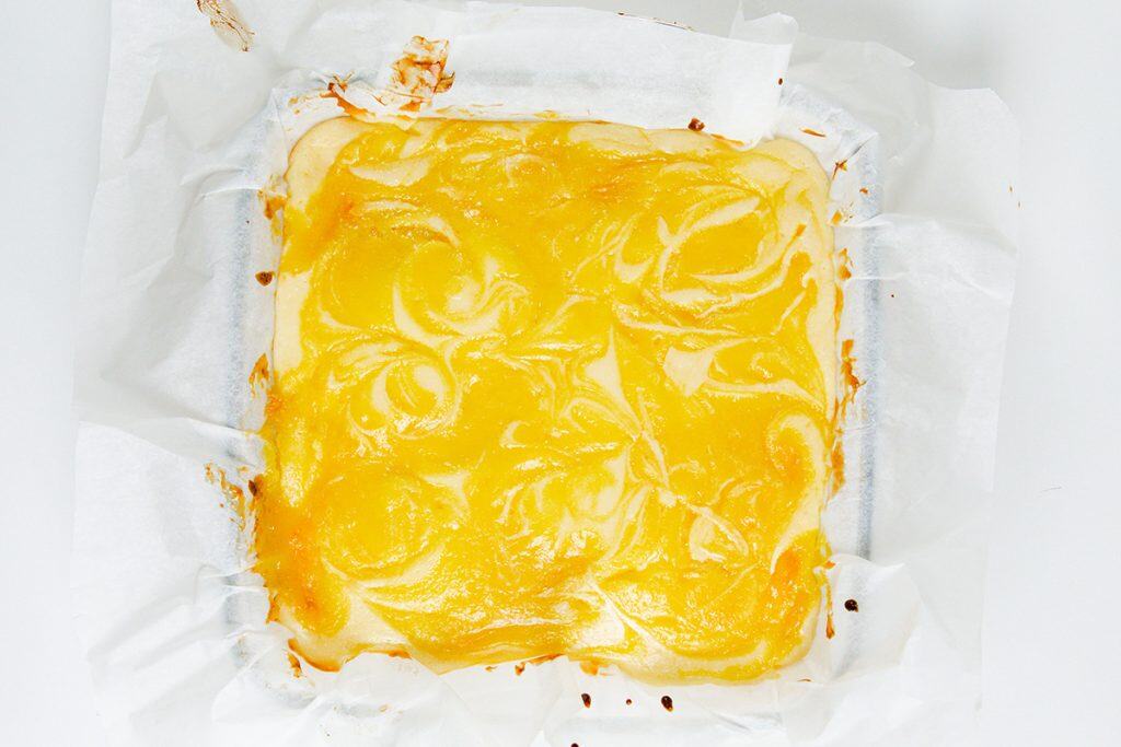 A square Lemon Cheesecake Blondie Bar dessert with a swirled yellow topping, baked in a parchment-lined pan, viewed from above on a white surface.