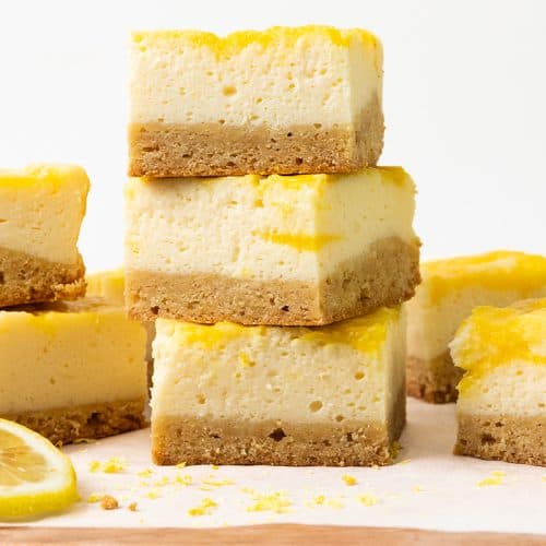 Stacked Lemon Cheesecake Bars with a crumbly crust and creamy yellow lemon topping, set on parchment with lemon slices and zest scattered nearby&mdash;perfectly blending classic blondie bars texture with zesty lemon cheesecake flavor.