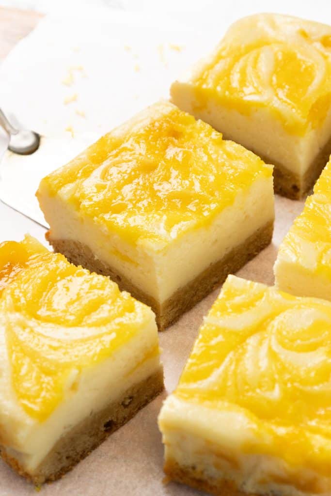 Four lemon cheesecake blondie bars with a golden yellow swirled topping are arranged on parchment paper. The bars have a creamy layer over a brown crust, and a spoon is partially visible in the background.