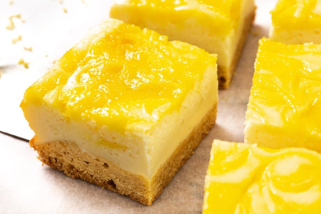 A close-up of Lemon Cheesecake Bars on parchment paper, featuring a golden brown crust, creamy cheesecake layer, and glossy yellow lemon topping with a swirled pattern.