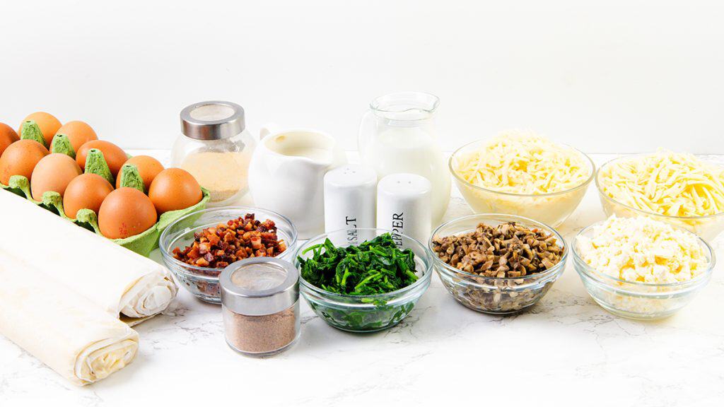 A selection of quiche ingredients for mini quiches&mdash;including eggs, puff pastry, cooked bacon, spinach, mushrooms, cheese, feta, milk, cream, and seasonings&mdash;arranged on a white surface for delicious bite-sized appetizers.