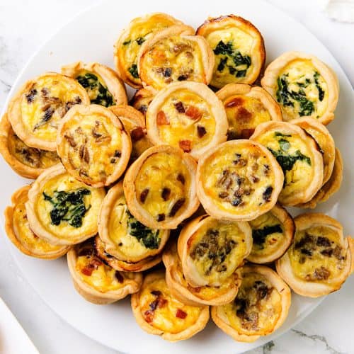 A white plate is piled with Mini Quiches featuring golden crusts and various fillings like spinach, cheese, and bacon, set on a white marble surface.