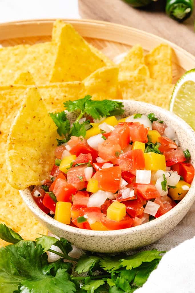 A bowl of fresh tomato salsa reminiscent of Pico de Gallo, with diced tomatoes, onions, yellow bell peppers, and cilantro, served with tortilla chips. Lime and cilantro garnish are on the side.