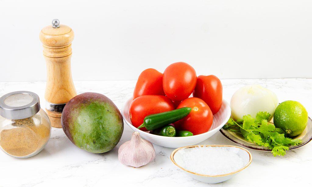 A variety of fresh ingredients for salsa or Pico de Gallo on a white surface, including tomatoes, jalapeños, mango, garlic, onion, lime, cilantro, a bowl of salt, ground seasoning, and a pepper grinder.