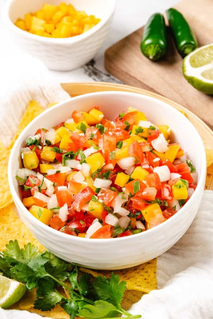 A white bowl filled with colorful mango salsa, a vibrant twist on Mexican salsa, made of diced mango, tomato, onion, cilantro, and jalapeño rests on a table next to fresh cilantro, lime, jalapeños, and tortilla chips.
