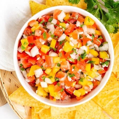 A bowl of colorful Pico de Gallo fresh salsa with diced tomatoes, yellow bell peppers, onions, and cilantro is surrounded by yellow tortilla chips on a wooden surface.