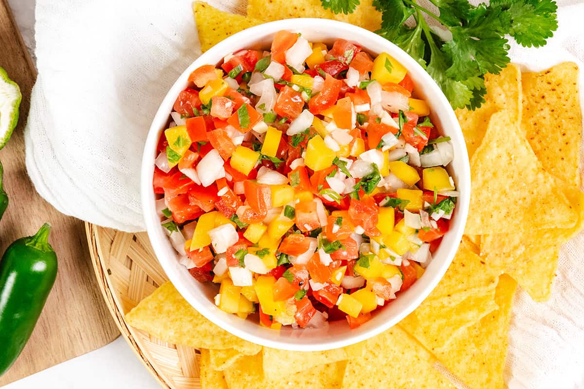 A bowl of colorful Pico de Gallo fresh salsa with diced tomatoes, yellow bell peppers, onions, and cilantro is surrounded by yellow tortilla chips on a wooden surface.
