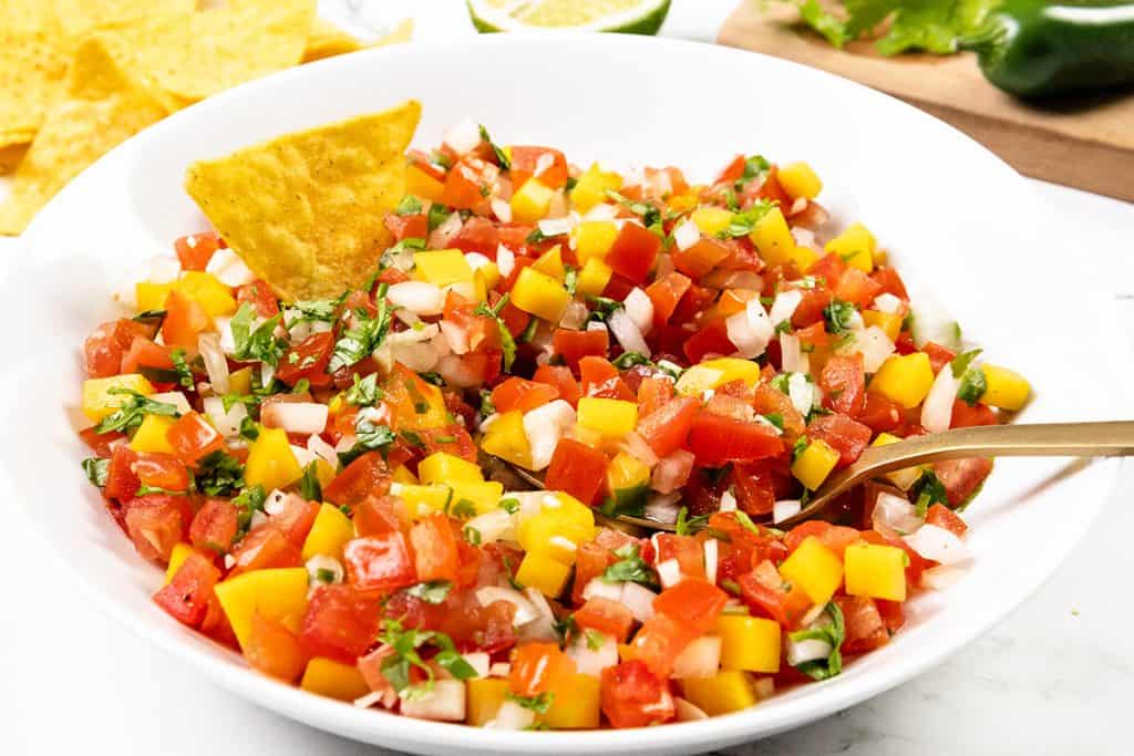 A white bowl filled with colorful Mexican salsa, featuring diced mango, tomatoes, onions, cilantro, and a tortilla chip dipped into the salsa. A spoon rests in the bowl, with chips visible in the background.