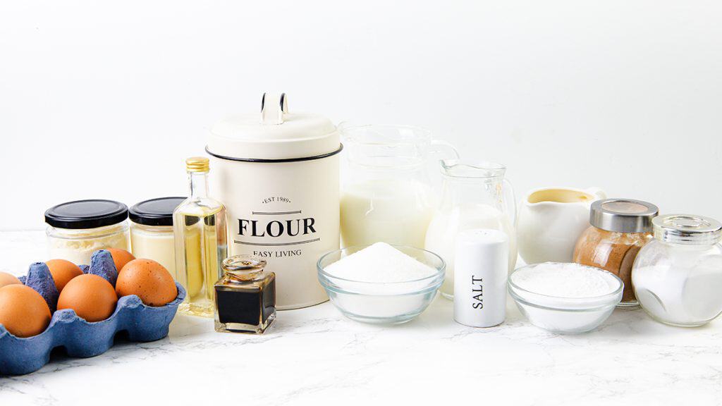 A collection of baking ingredients on a white surface, perfect for making Tres Leches Cake, including eggs, flour, sugar, milk, vanilla extract, oil, salt, cinnamon, baking powder, and a flour canister.