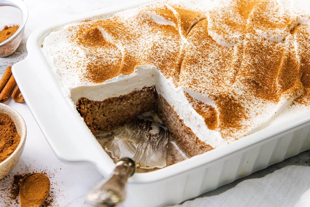 A rectangular white baking dish filled with Tres Leches Cake, its cinnamon-topped surface covered in whipped cream. A slice has been served, revealing the moist, brown cake inside. A serving utensil rests in the dish. Bowls of cinnamon are nearby.