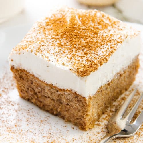 A square slice of Tres Leches Cake with whipped cream topping, dusted with cinnamon, sits on a white plate beside a fork.
