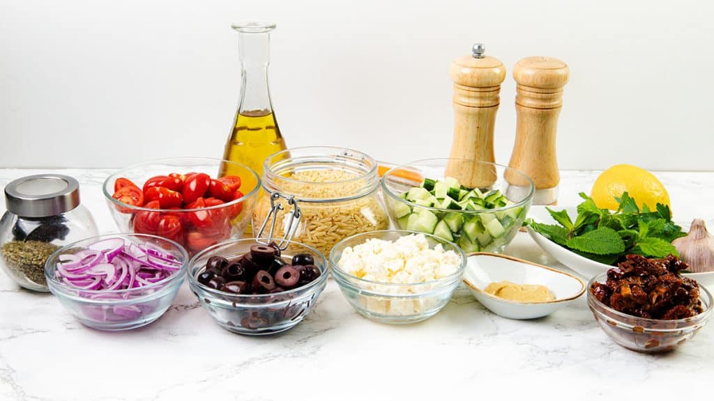 A variety of Mediterranean salad ingredients are arranged on a white surface, showcasing a classic Greek Orzo Pasta Salad with red onions, olives, cherry tomatoes, feta cheese, cucumber, dried tomatoes, olive oil, herbs, pepper, salt, lemon, garlic, and dressing.