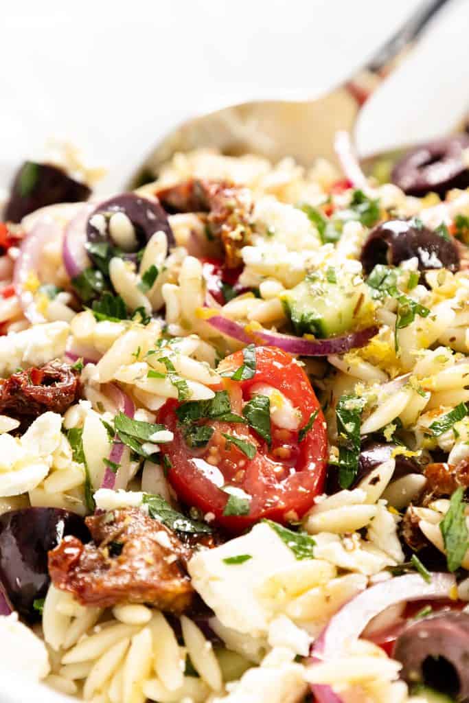 A close-up of a Greek Orzo Pasta Salad with cherry tomatoes, Kalamata olives, red onion, feta cheese, sun-dried tomatoes, and fresh herbs, with a serving spoon in the background.