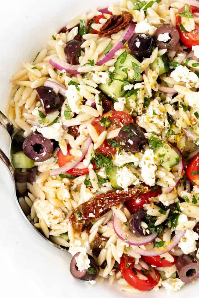 A close-up of a vibrant Greek Orzo Pasta Salad with Kalamata olives, cherry tomatoes, red onions, feta cheese, cucumber, sun-dried tomatoes, and fresh herbs, served in a white bowl with a spoon.