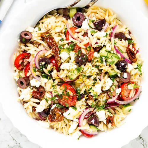 A white bowl filled with Greek Orzo pasta salad, topped with cherry tomatoes, Kalamata olives, red onion, sun-dried tomatoes, feta cheese, and chopped herbs. A serving spoon is in the bowl. Lemons and parsley are nearby.